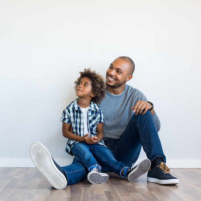 African father and his son sitting on floor and looking up in a blank wall. Happy dad and little boy sitting in an empty room. Young black man with his child thinking and pensive with copy space.; Shutterstock ID 692844577