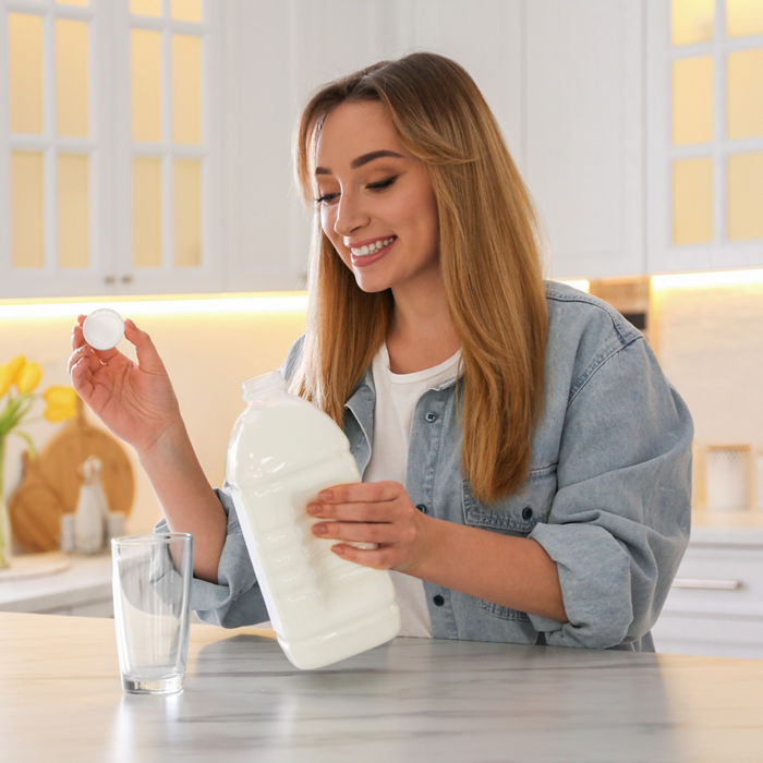 Young woman pouring milk from gallon bottle into glass at white marble table in kitchen; Shutterstock ID 2208427439; purchase_order: POL website_OmyaPET; job: ; client: ; other: 