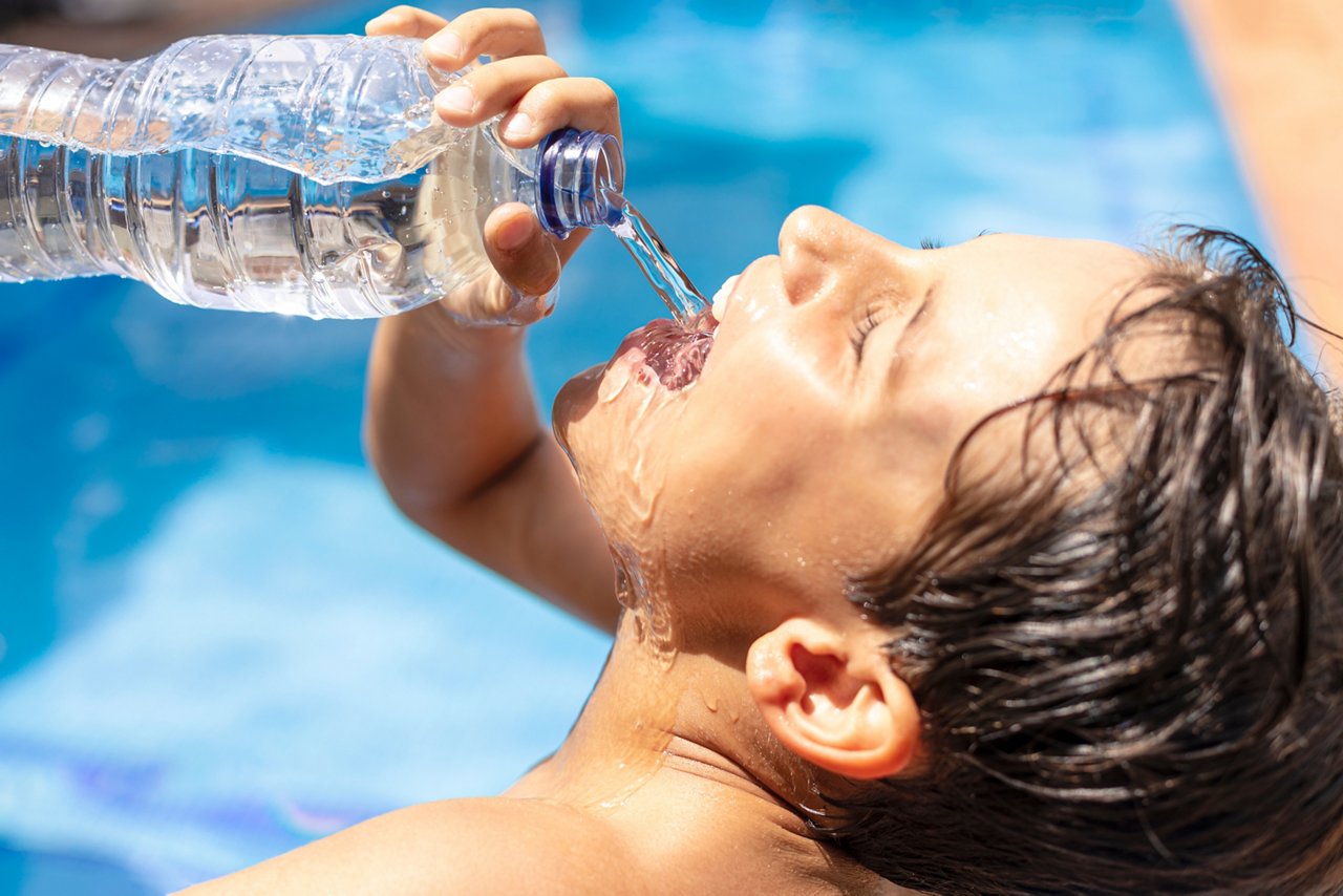 Boy drinking water from a plastic bottle in in the pool on a hot summer day. Young man drinking a lot of water after sports to avoid heat stroke. Child drinking plenty of water after physical exercise