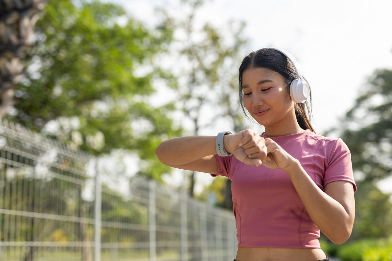 Asian women in sportswear checking smartwatch while listening to music with headphones during outdoor workout, representing fitness tracking, healthy lifestyle, wearable technology.; Shutterstock ID 2724808897; purchase_order: OPP Website & Collateral; job: ; client: ; other: 